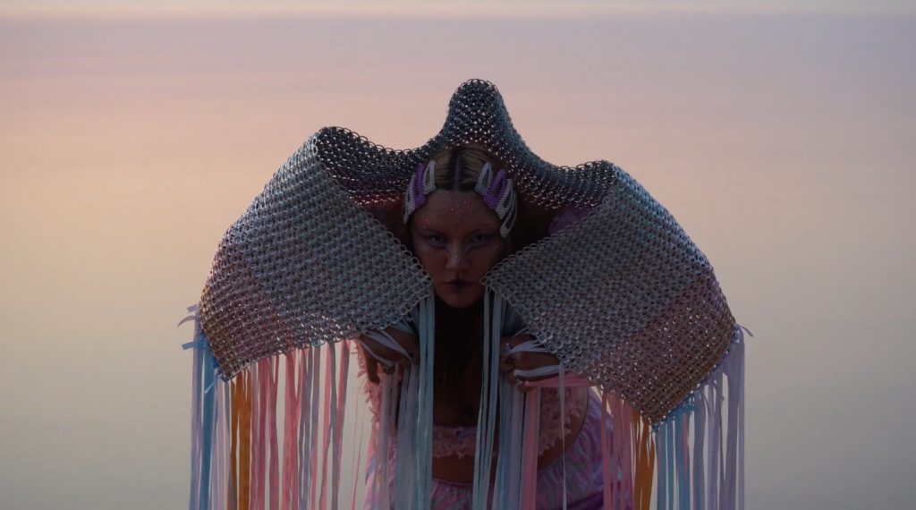 Image of a person leaning forward in front of a pink and bluish background, with a fringed shawl over their shoulders.