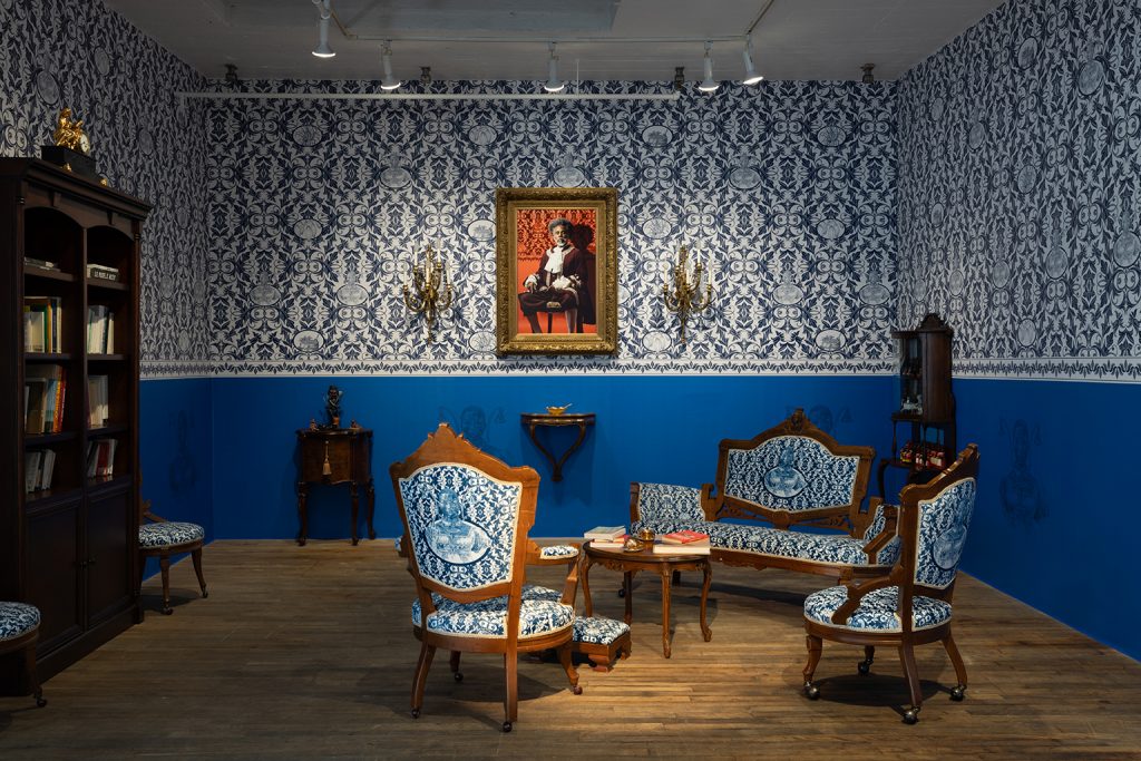 Une pièce avec un papier peint à motifs bleus et blancs, des meubles rembourrés assortis, une étagère et un portrait encadré au mur. La pièce est dotée d'un sol en bois et de divers objets de décoration. A room with blue and white patterned wallpaper, matching upholstered furniture, a bookshelf, and a framed portrait on the wall. The room has wooden flooring and various decorative items.