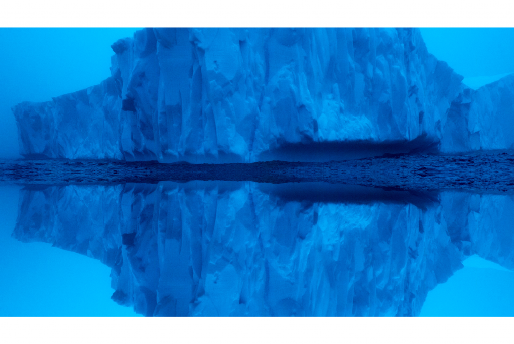 Un iceberg massif se profile au-dessus de l'eau, se reflétant parfaitement dans la surface sereine qui l'entoure. A massive iceberg looms over the water, perfectly mirrored in the serene surface beneath it.