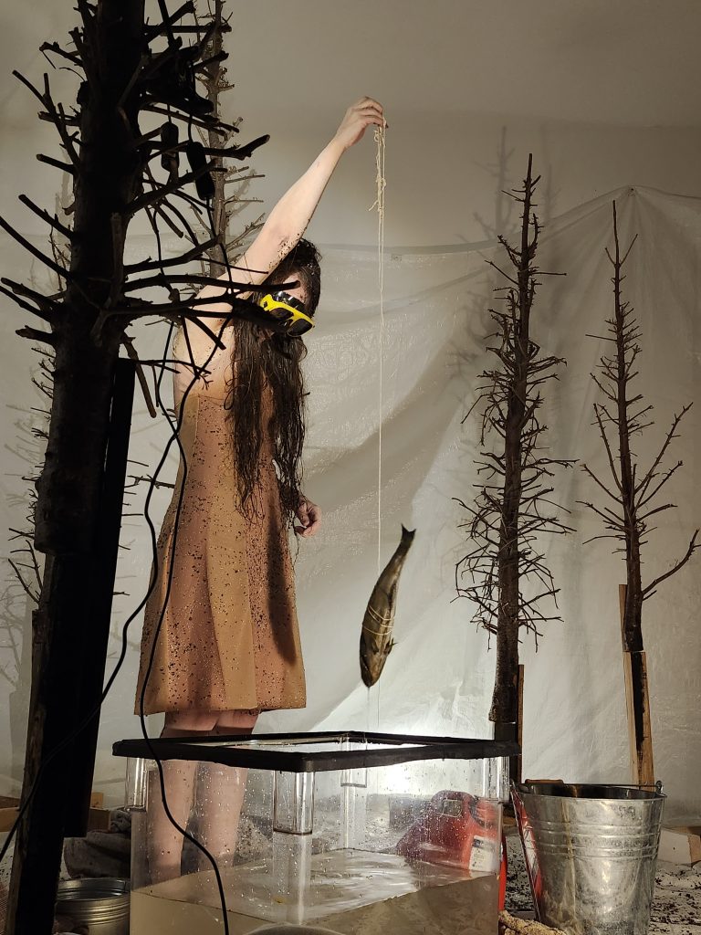 A woman with long, damp brown hair, partially covered in dirt, stands in an immersive art installation. She wears a beige dress speckled with dirt and yellow safety goggles. Her arm is raised, holding a string from which a fish is suspended above an aquarium. Around her, bare, dark trees with dry branches create an enigmatic atmosphere. The background consists of a stretched white fabric, giving the scene a theatrical impression.