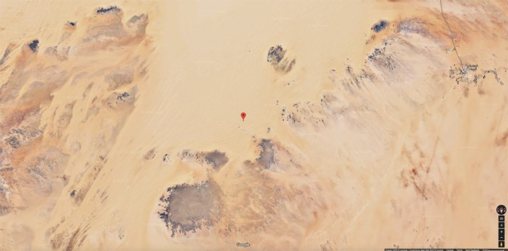 Une vue satellite d'un paysage désertique avec une épingle rouge marquant un point spécifique au milieu d'une étendue de sable. Le terrain est composé de dunes de sable et de formations rocheuses aux teintes de beige, marron et noir. Un petit village est visible dans le coin supérieur droit, relié par une route. L'image provient de Google Maps.