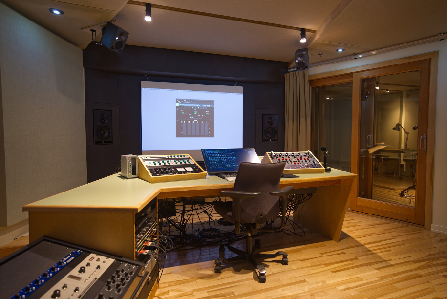 Un studio d'enregistrement professionnel équipé d'une grande console de mixage, d'un écran d'ordinateur, de haut-parleurs et d'une vitre insonorisée donnant sur une cabine d'enregistrement.